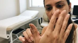 Mão feminina observa atentamente mancha escura longitudinal na unha do polegar em ambiente clínico.